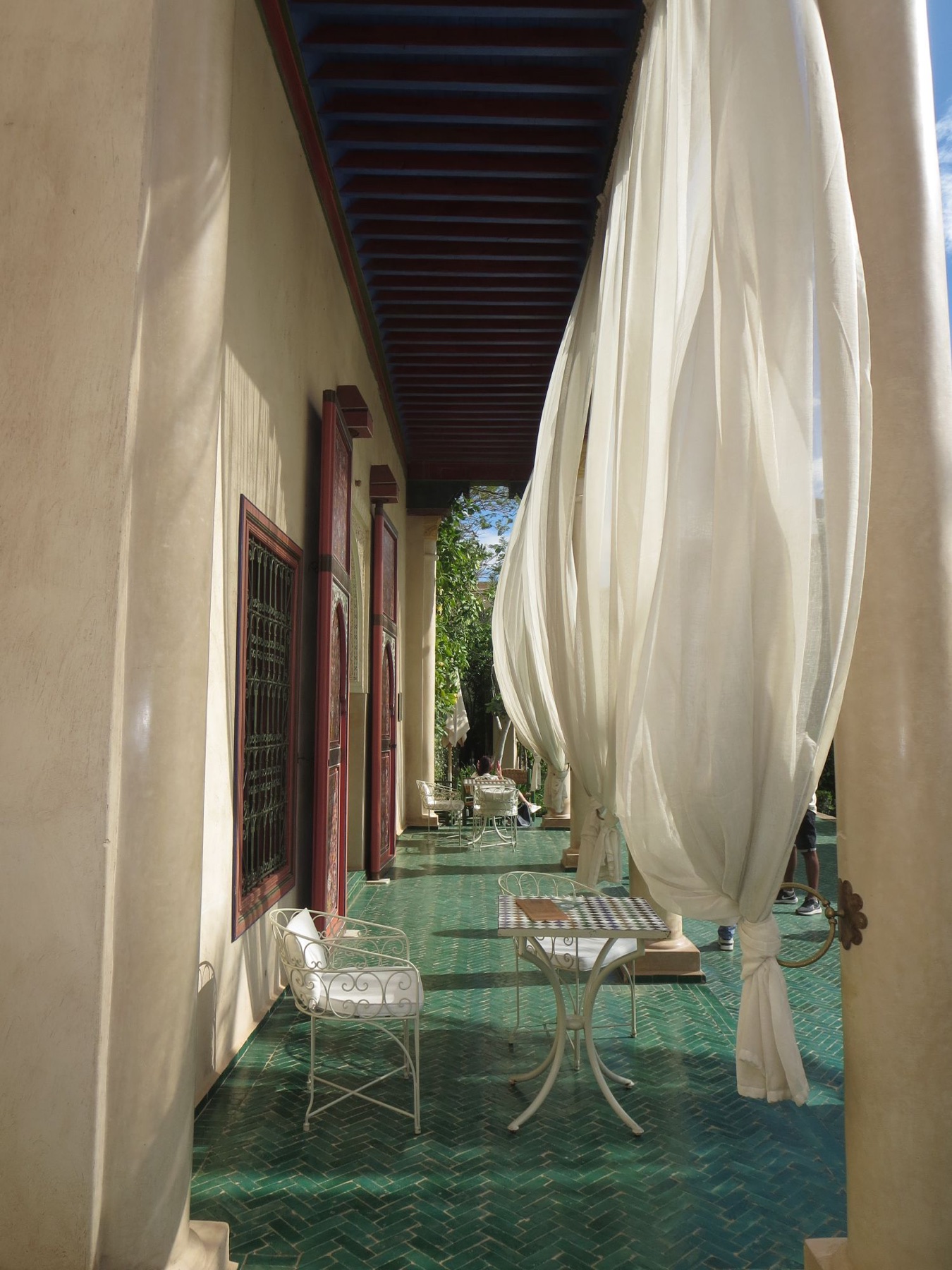 A sunlit veranda with tall white curtains moving in the breeze, green tiled floor and white iron chairs